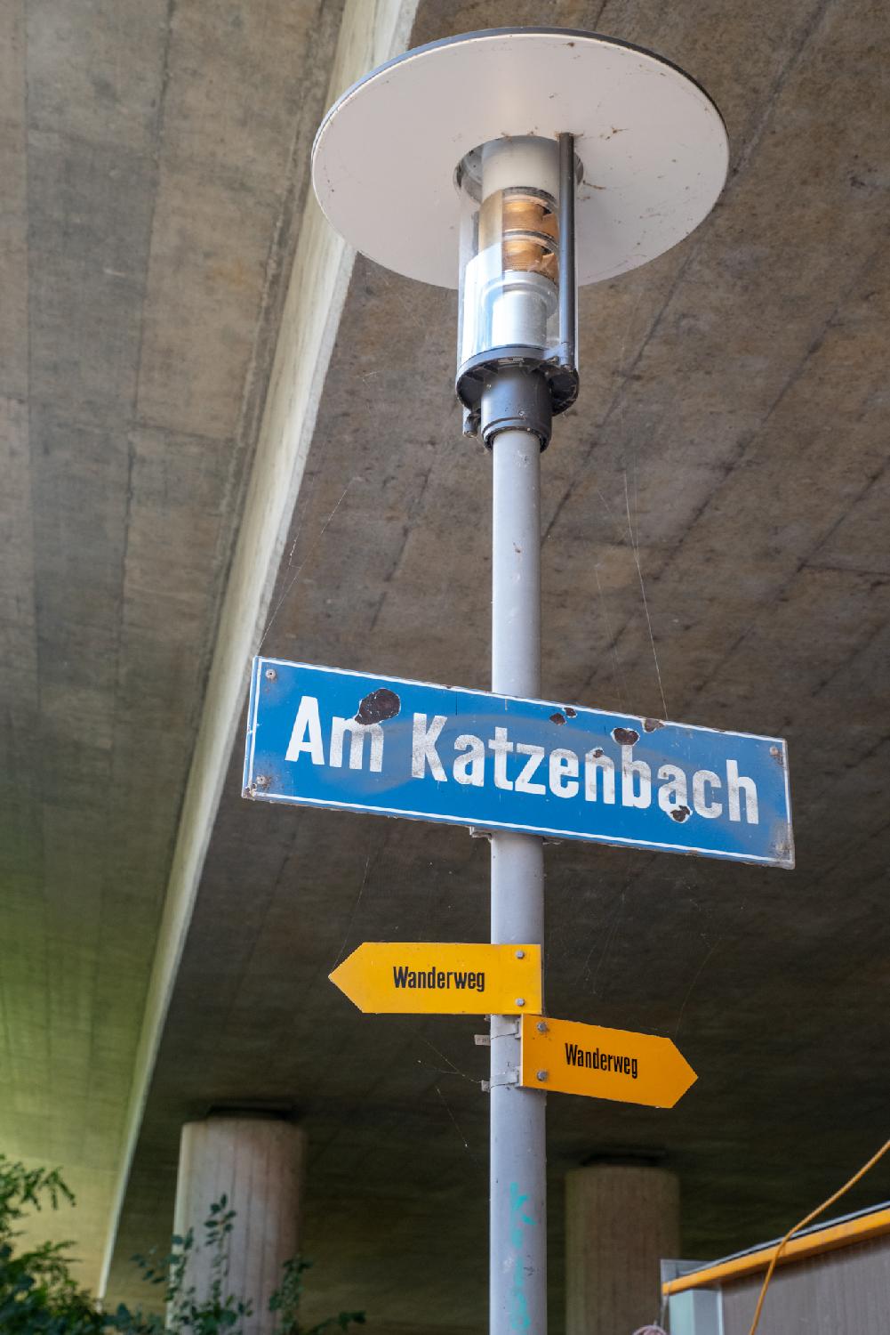 A street light under a concrete bridge, with a "Am Katzenbach" street sign (covered in spider webs) and a couple of yellow hiking path (Wanderweg) signs.