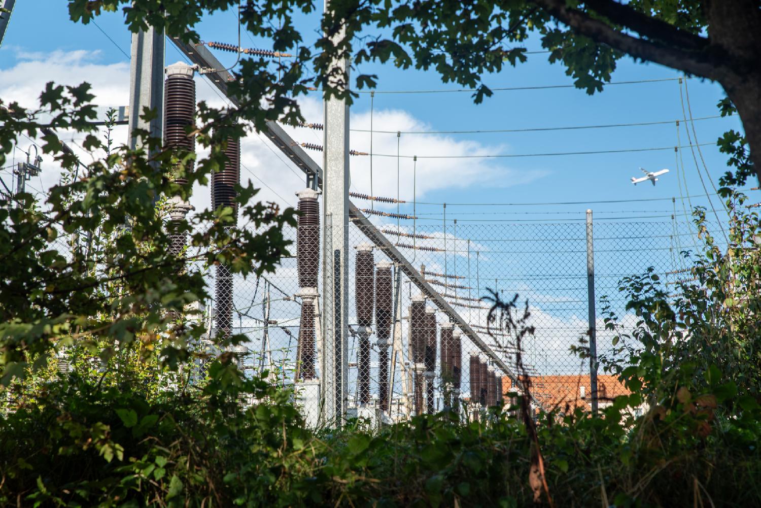 Seebach Unterwerk in Zürich: an electrical substation with pylons and coils, between trees. A plane is taking off in the background.