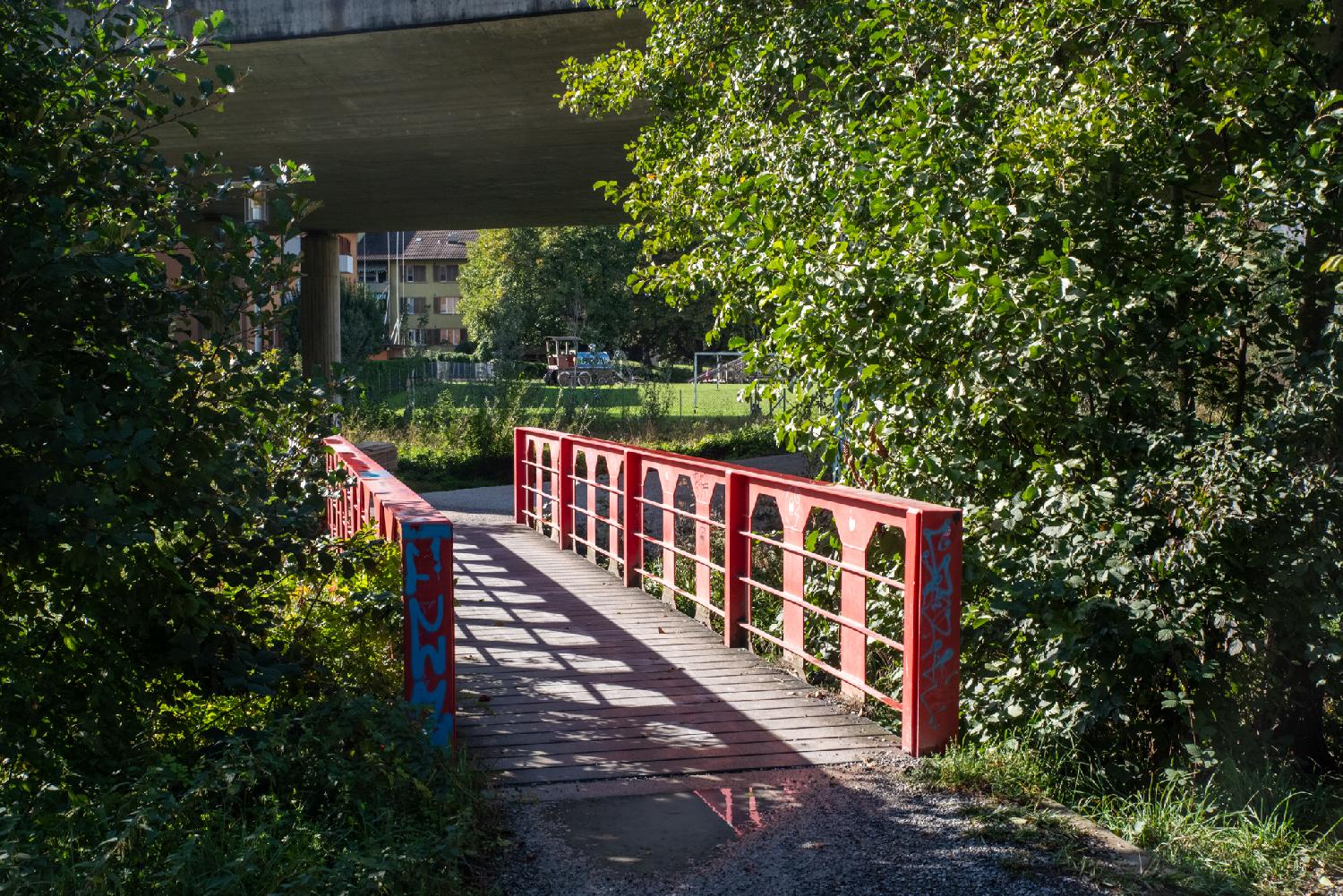 A wooden bridge with red metallic beams between trees. Another larger concrete bridge is visible on top of the picture.