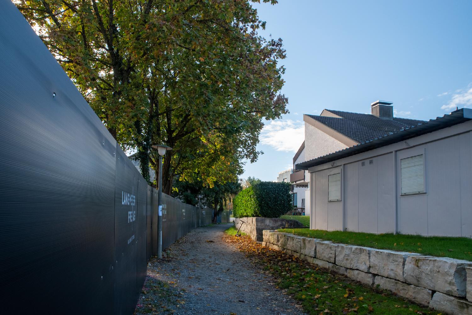 A gravel path with, on the left side, black construction fence (with trees behind it) and, on the right side, 1-2 story habitation buildings.