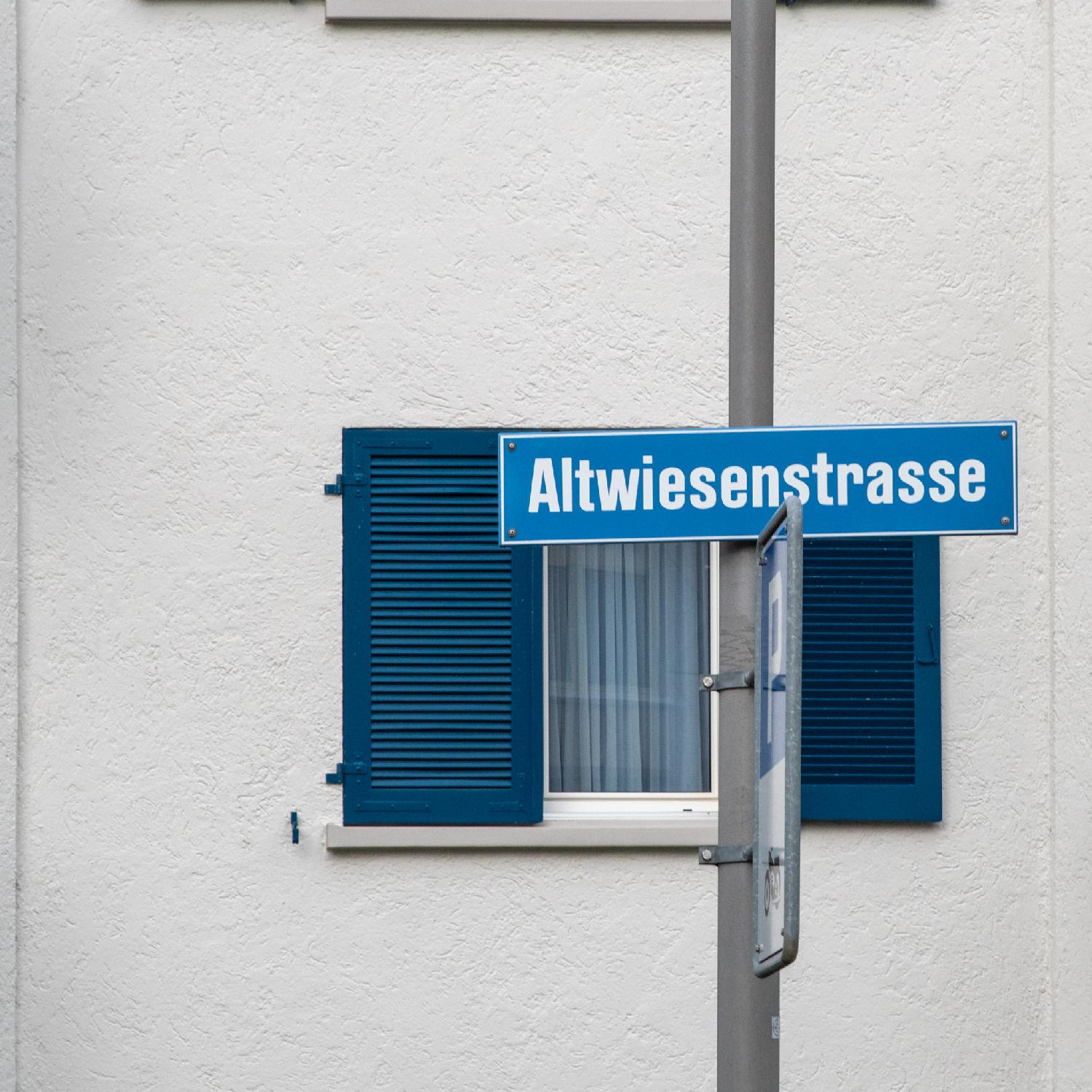 Street name sign for Altwiesenstrasse in Zürich: a blue street sign on a metallic pole, in front of a white wall and a window with blue blinds.