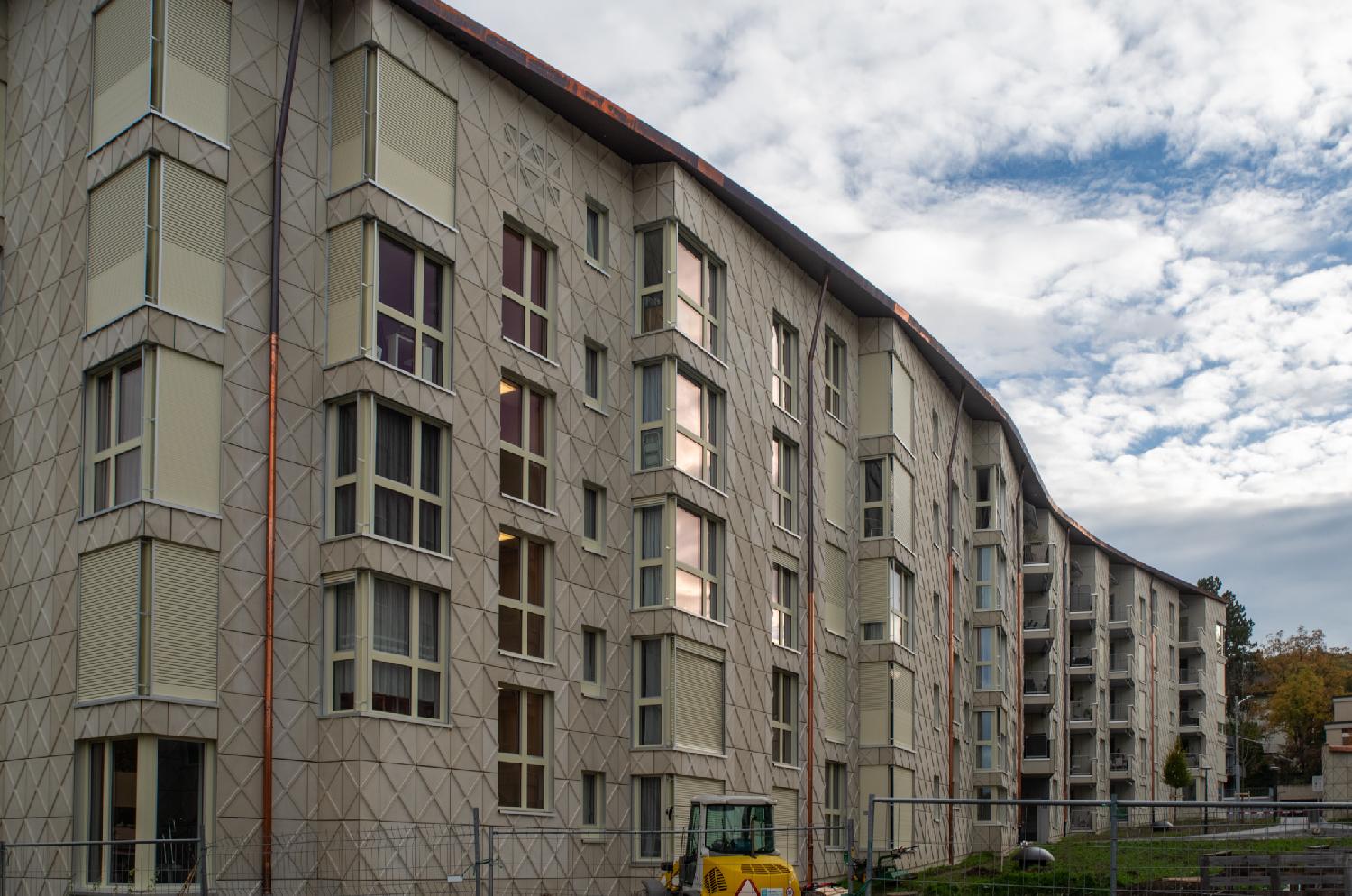 4-story beige buildings in a wavy alignment. The buildings are new, there's still a small excavator and works fences around it.