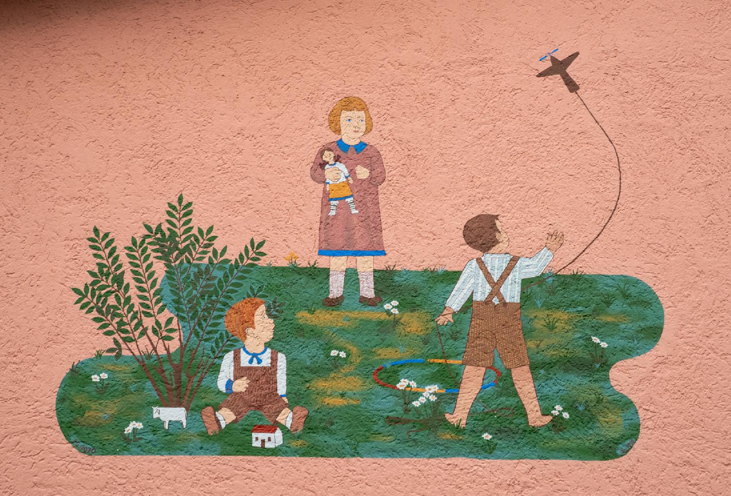 Painting on a wall on Altwiesenstrasse in Zürich: three children playing in a grean meadow and looking at an airplane, painted over a pink house wall