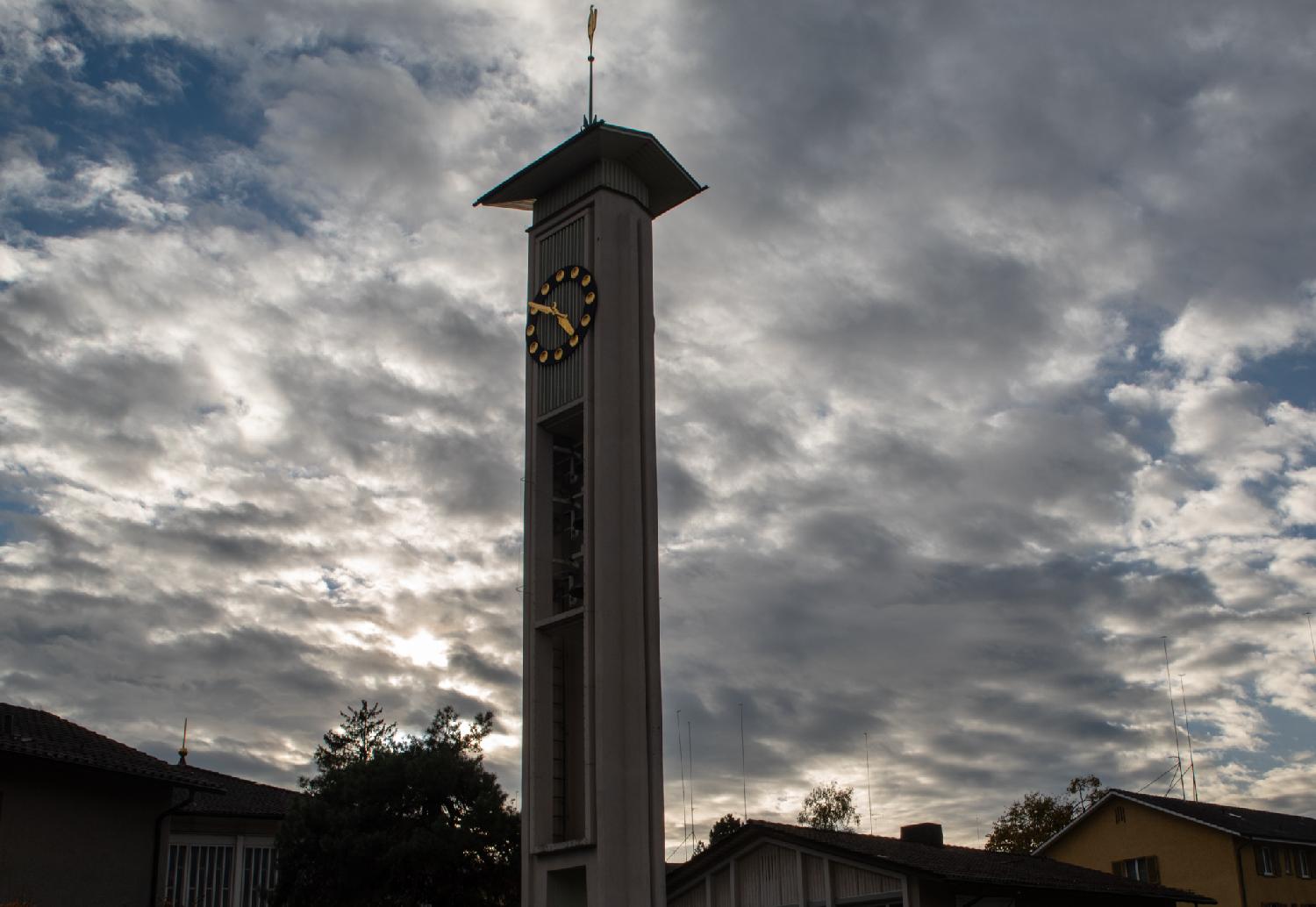 Stefanskirche Hirzenbach on Altwiesenstrasse in Zürich: a church tower with three bells, a golden clock and a golden weathercock, contre-jour against a cloudy sky