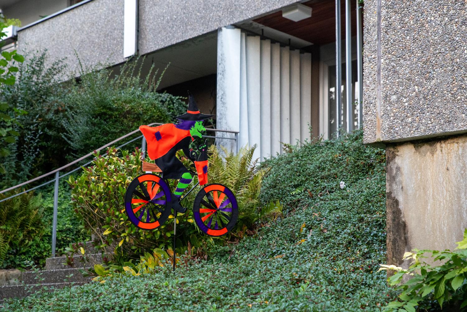 Halloween decoration in front of a building door: a green witch with a red cape riding a bike, on top of a bed of ivy