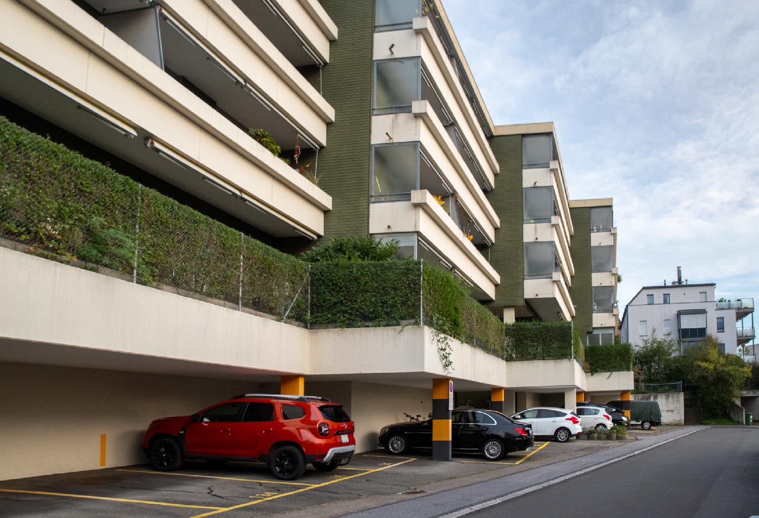 A block of four 3-story buildings above parking lots (with cars parked in it). The floor above the parking lot has green hedges.