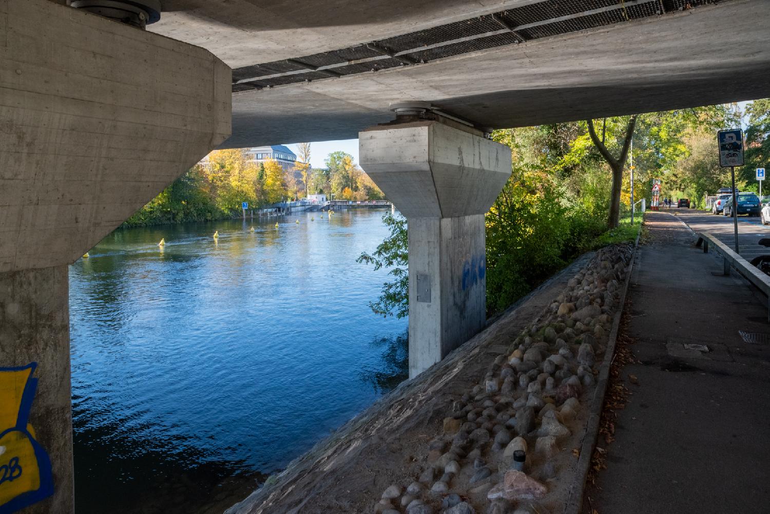 On the left of the picture, the Limmat river (blue and calm), on the right of the picture a narrow street; a small slope with large pebbles inbetween, and the bottom of a bridge on top.