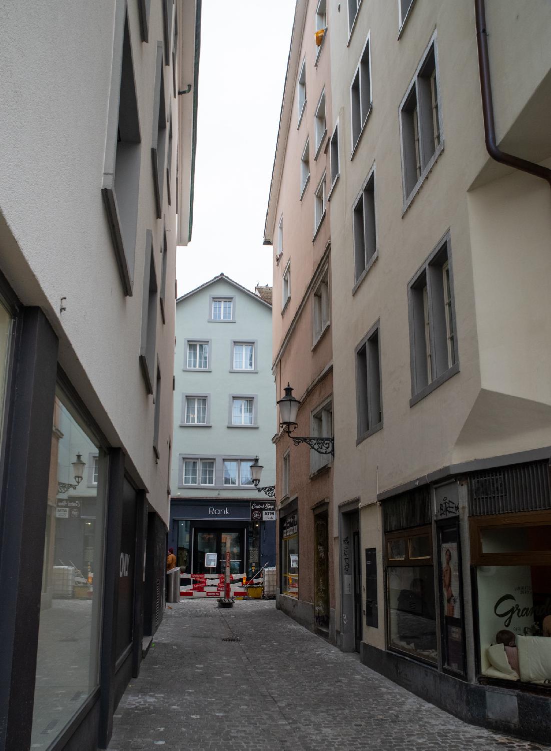 Am Rank in Zürich, direction Niederdorfstrasse: a narrow paved alley between two 5-story building rows, and another light green 5 story-building visible in the street perpendicular to it. Said street has construction work.