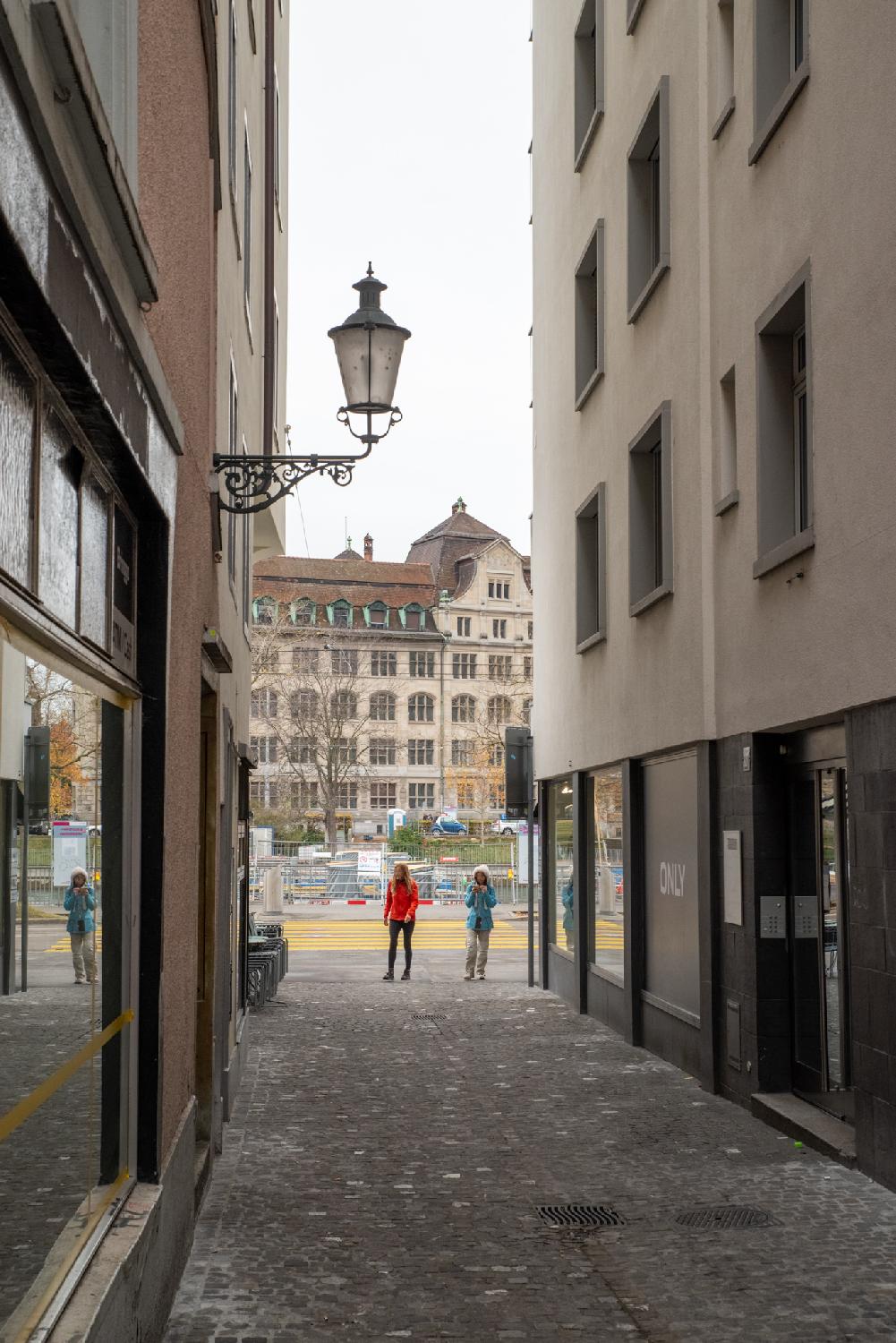 Am Rank in Zürich, direction Niederdorfstrasse: a narrow paved alley between two building rows, with a larger 5-story administrative building visible in the background.
