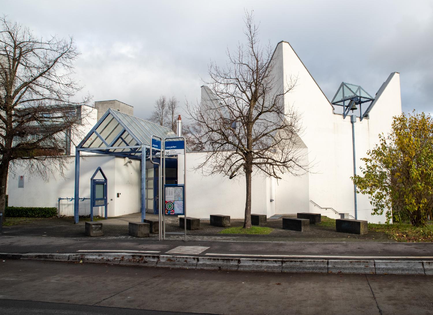 Kirche Suteracher: a white building behind a bus stop. The building has a bell under a glass roof; the building entrance also has a glass roof. The building looks kind of like a crown with four high points.