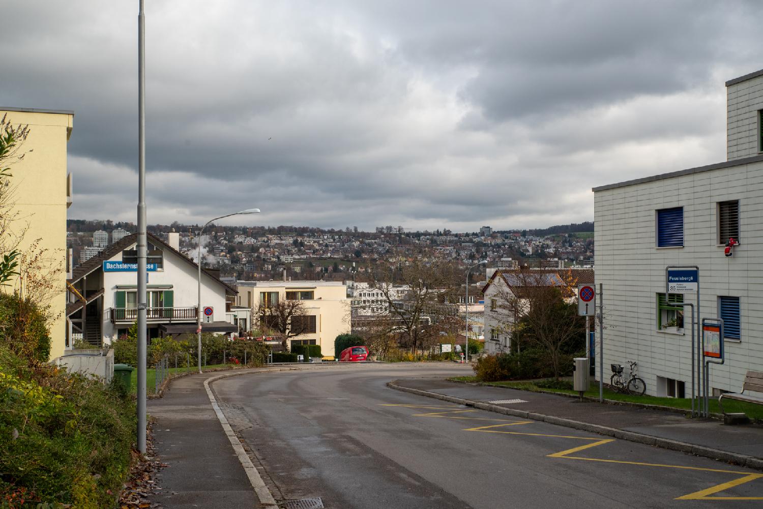 A residential street turning right, with a bus stop on the right, and dense urban area visible in the background