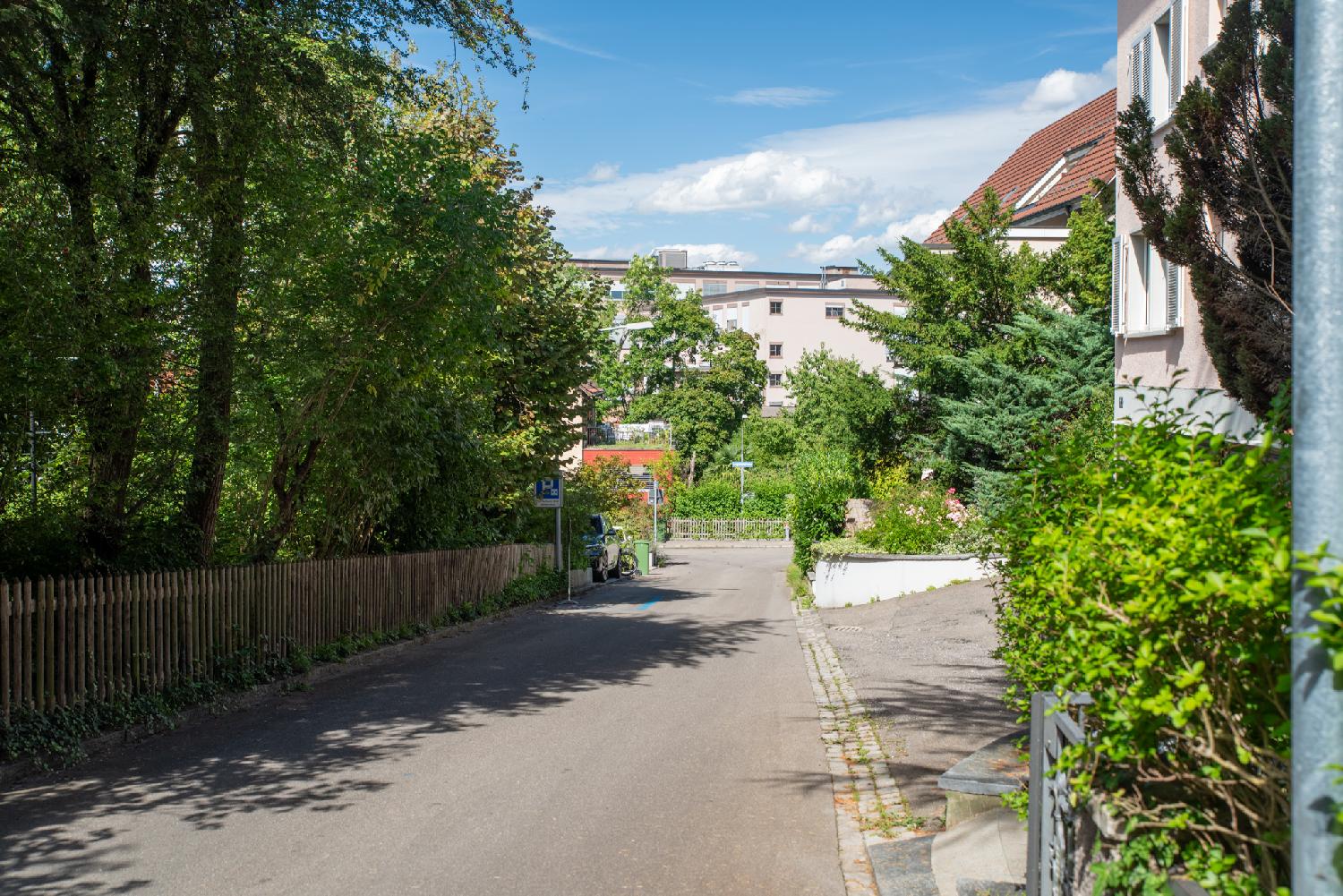 A narrow street with a wooden fence and trees on the left, and 2-3-story residential buildings on the right, and more buildings on the perpendicular street in the background.