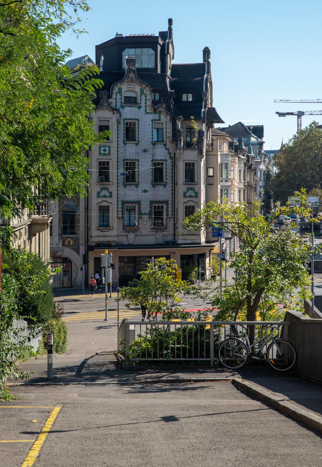 A narrow street that ends in a footpath. On the perpendicular street, a building decorated with white and green tiles and curved structures.