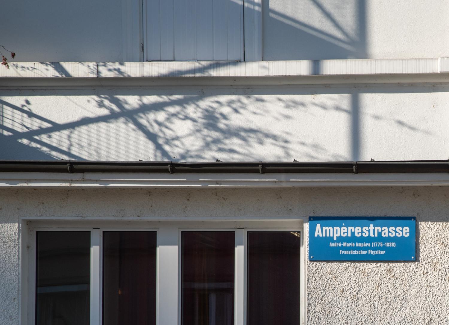 Blue sign on a white wall next to windows. The sign reads "Ampèrestrasse - André-Marie Ampère (1776-1836) - Französischer Physiker".