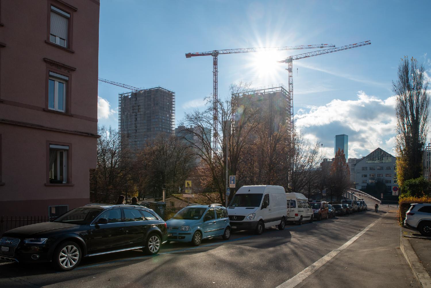 A small road ending on a bridge, with a lot of cars parked on the left of it. In the background, there are buildings in construction and a glass building (the Prime Tower).