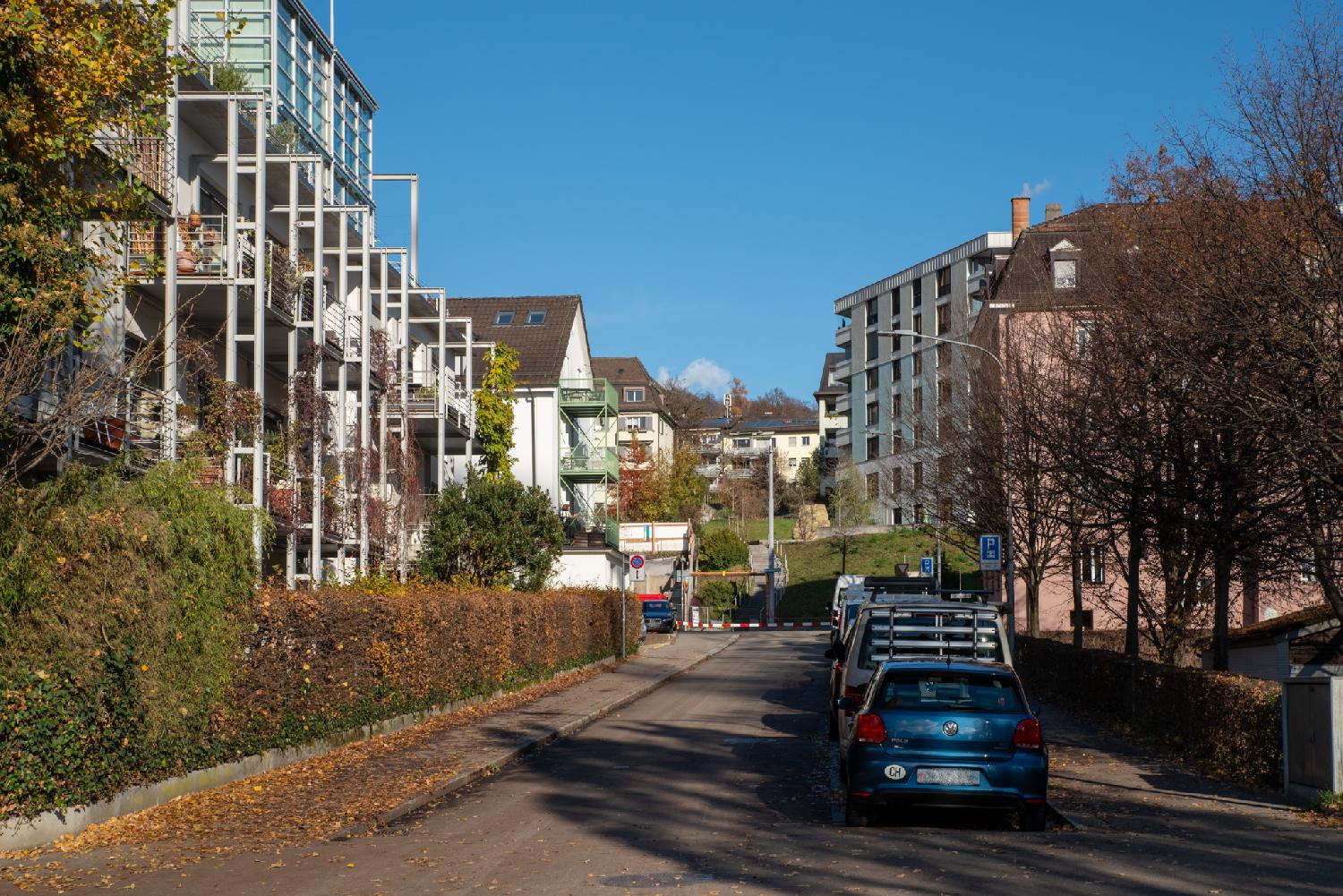 A narrow street with brown/orange hedges on each side, and 3-4-story residential buildings on each side, as well as a row of cars parked on the right.