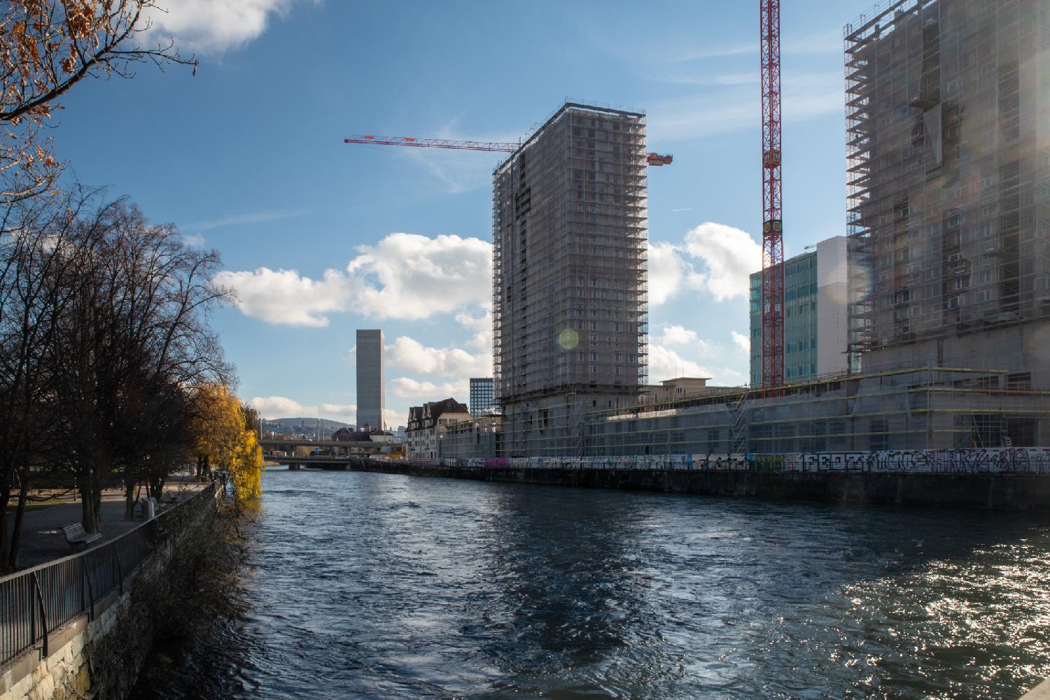 A river, seen from a bridge, with 5-6-story buildings on one bank, trees on the other bank, and a tiny island in the middle.