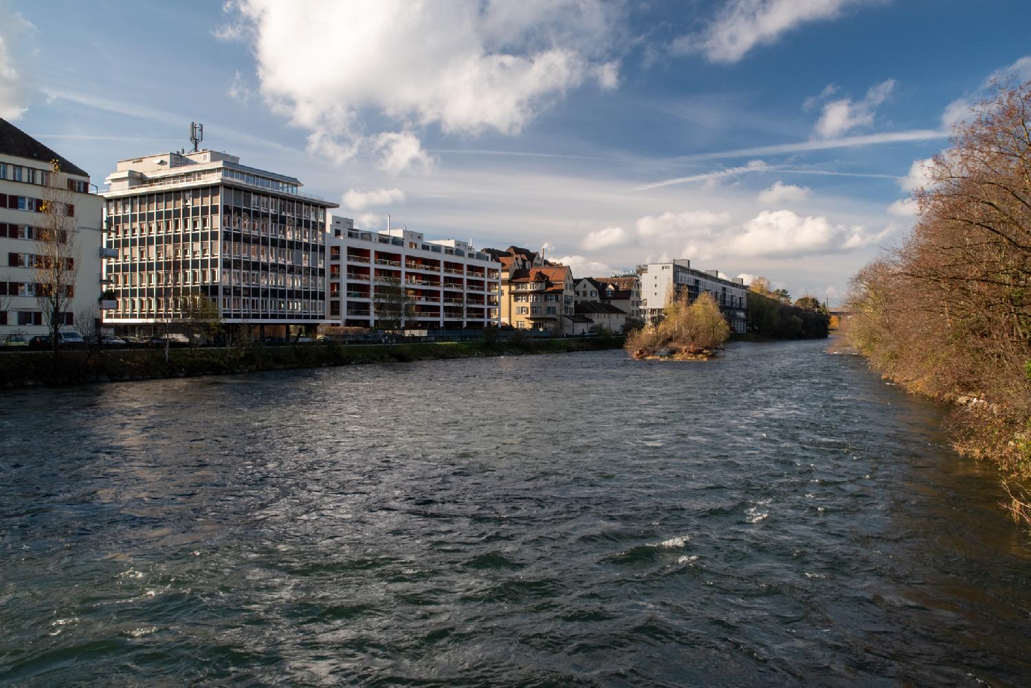 A river, seen from a bridge, with 5-6-story buildings on one bank, trees on the other bank, and a tiny island in the middle.