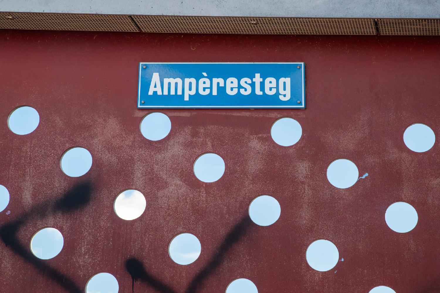 A blue "Ampèresteg" street sign riveted on a red metallic wall with holes.