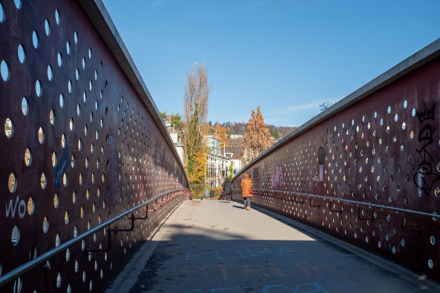 A pedestrian bridge with, on each side, high metallic walls with holes. There's graffiti on the walls and on the ground. The background has a few buildings and trees.