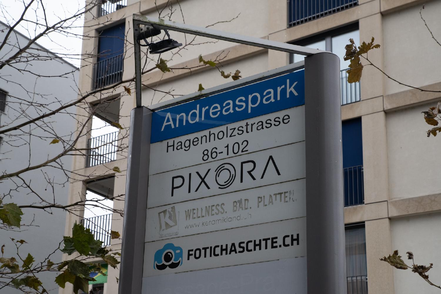 A blue "Andreaspark" sign, with more signs below it (Hagenholzstrasse 86-102, and some local business signs), in front of a beige building.