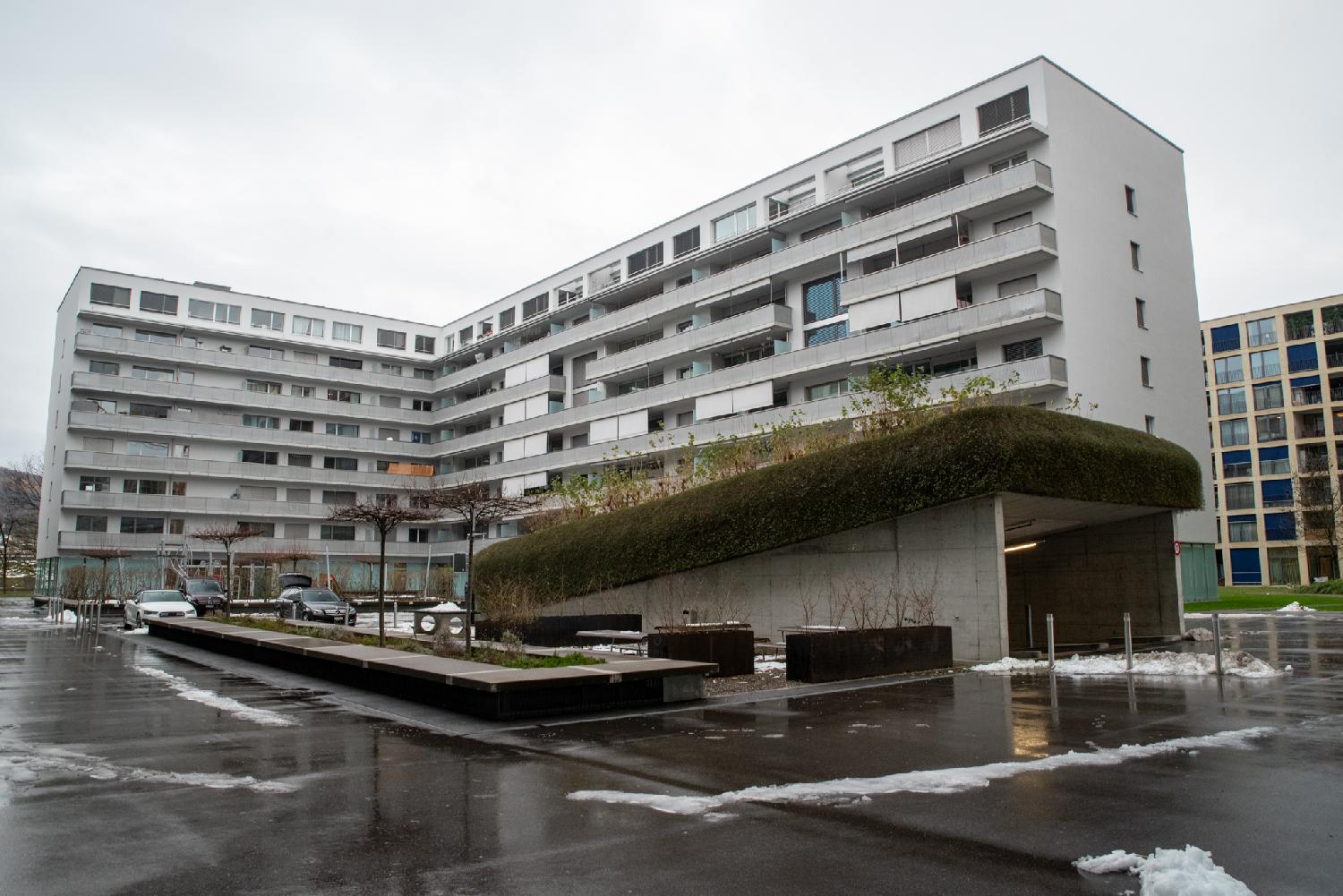 A L-shaped 8-story residential building in the background, with a large underground parking lot entrance in the foreground. There's a bit of snow on the ground.