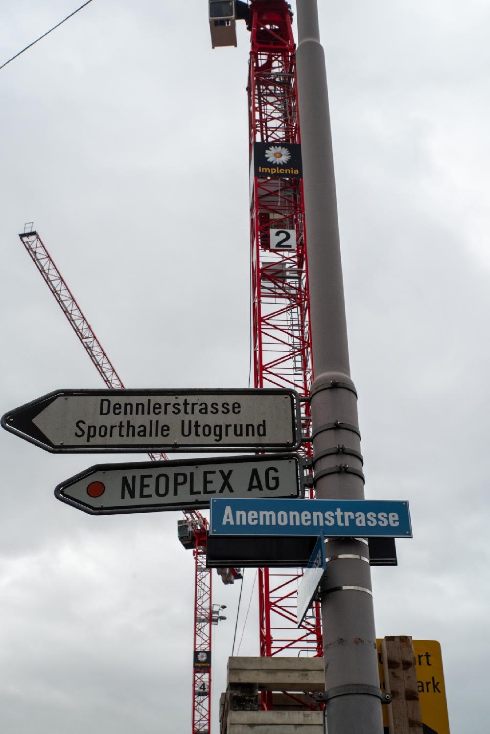 A blue "Anemonenstrasse" on a high grey pole in front of a red crane. There are also arrows to Dennlerstrasse, Sporthalle Utogrund, and Neoplex AG, pointing to the right.
