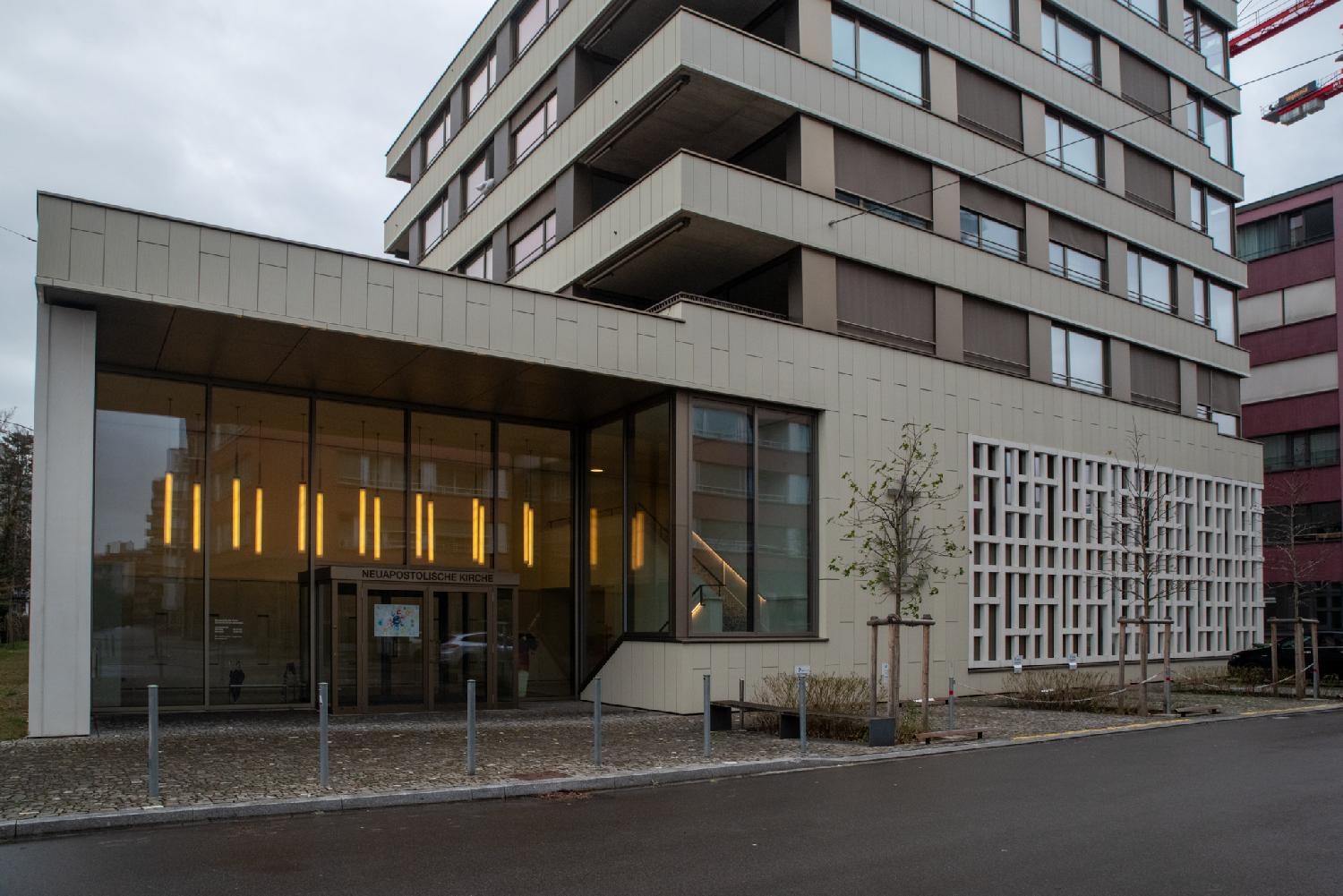 An L-shaped building that has, on the ground floor, a sign for "Neuapostolische Kirche" and small decorative windows and, on the higher floors, what looks like an apartment building.