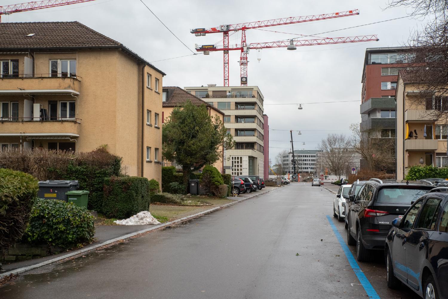 A two-way street with cars parked on each side and 4-to-10-story building on each side. There are red cranes in the background.