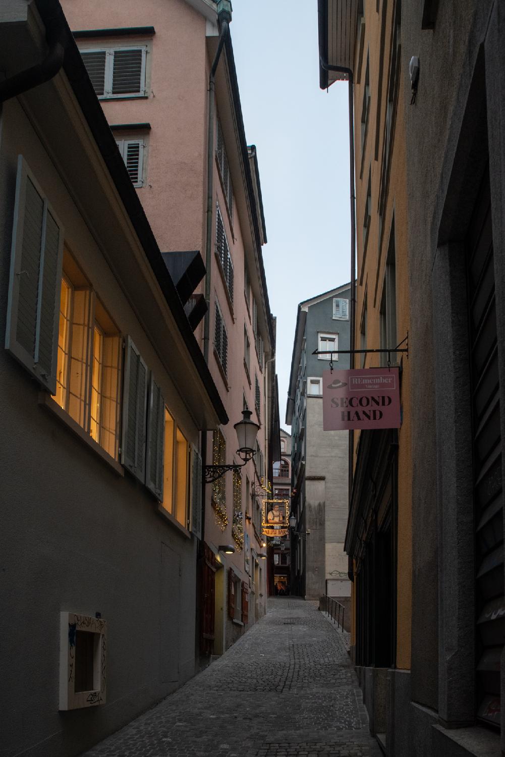 A narrow paved alley between 5-6 storey buildings. A decorated shop hanging sign is visible in the end of the street.