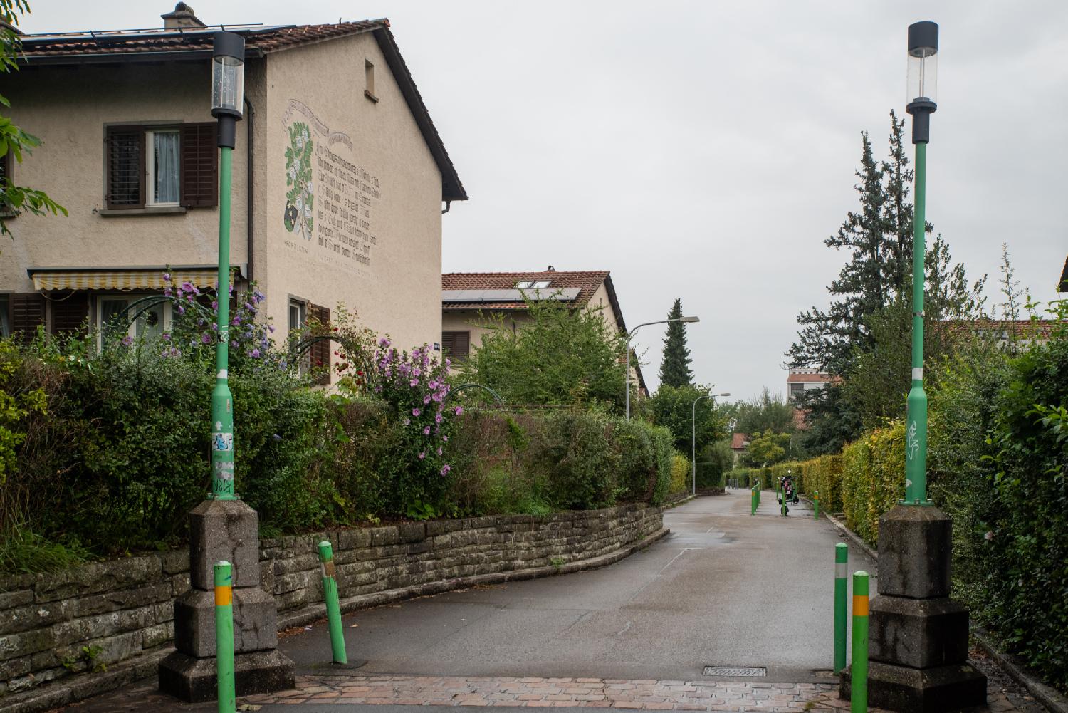 A residential street with 2-storey buildings and hedges on both sides of the street. The forefront building has an (unreadable) large relief inscription on one of the walls.