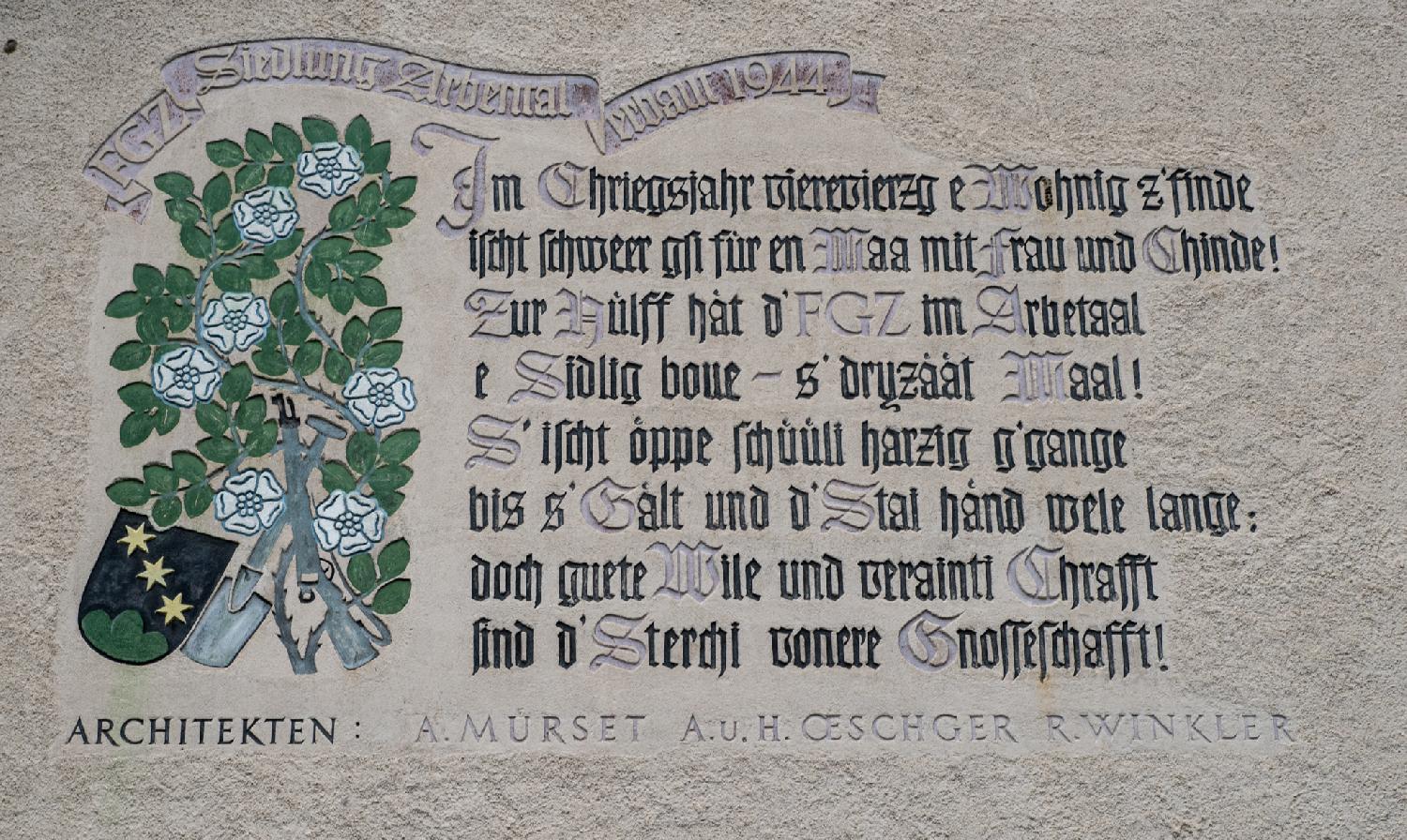 Relief on a wall with in image of white flowers and green leaves, as well as tools including a shovel, and a coat of arms with three stars and greenery. The text of the inscription is available in the caption of the image.