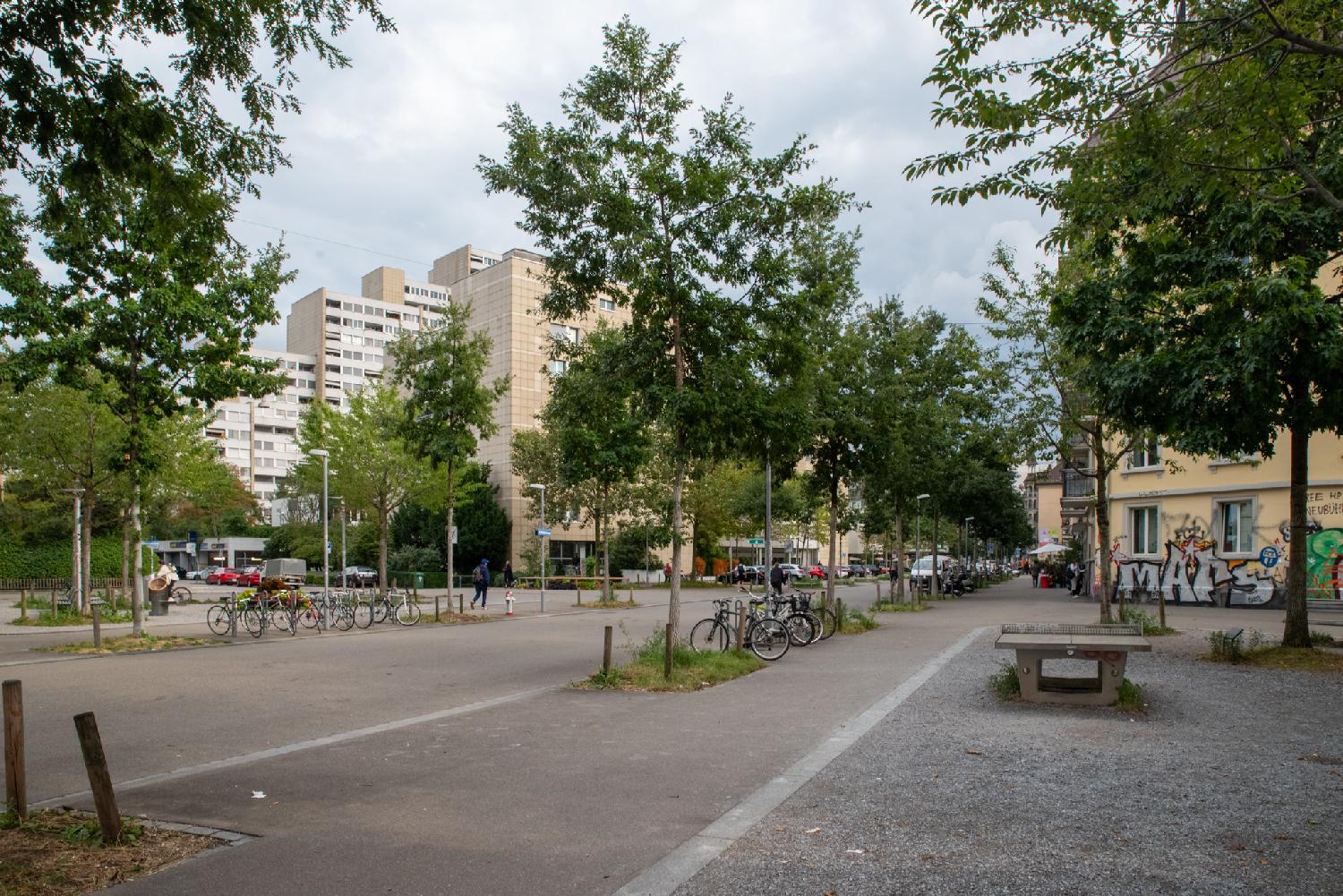 A square with trees, a concrete ping-pong table and bikes, between residential buildings (high-rises in the background, lower-rises closer)