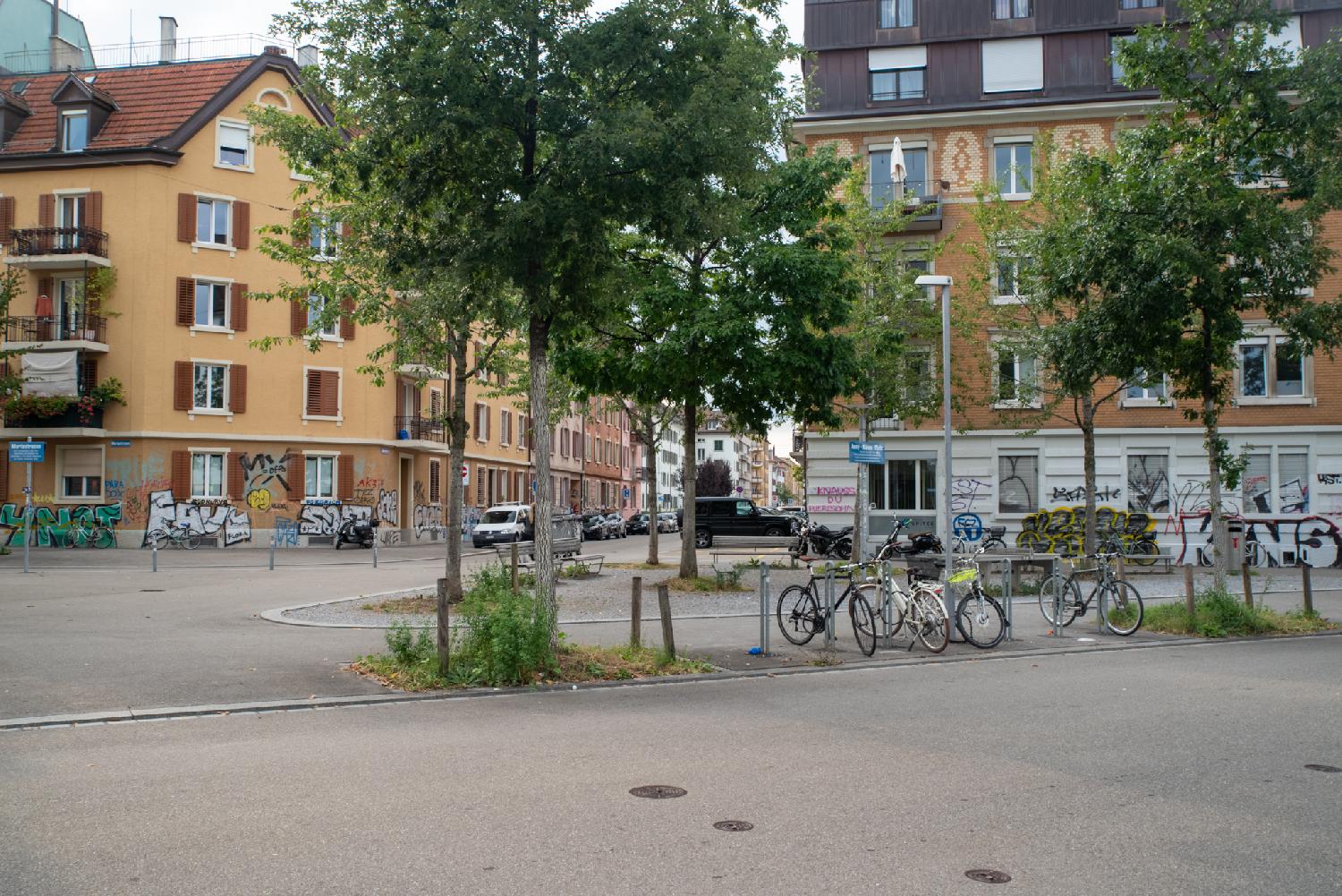 A square with trees and bikes in front of 5-story residential buildings with a lot of graffiti