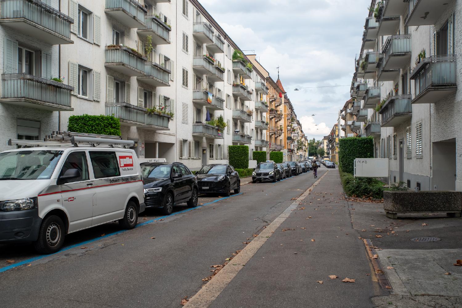 A residential street with 5-storey buildings on each side and cars parked on the left side.