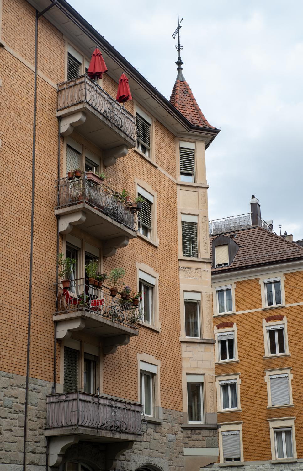 A brown 5-storey building with protruding bay windows on the corner and balconies on the façade. The bay window part have a small spire and a weathervane on top.