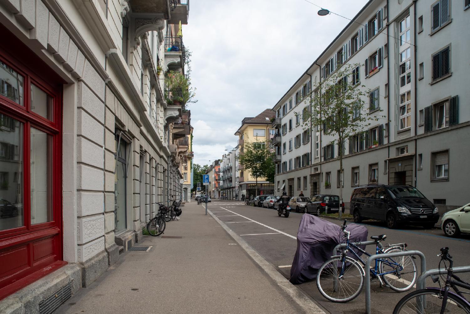 A residential street with 5-storey buildings on each side, empty parking spots and a few bikes on the left, and cars parked on the right.