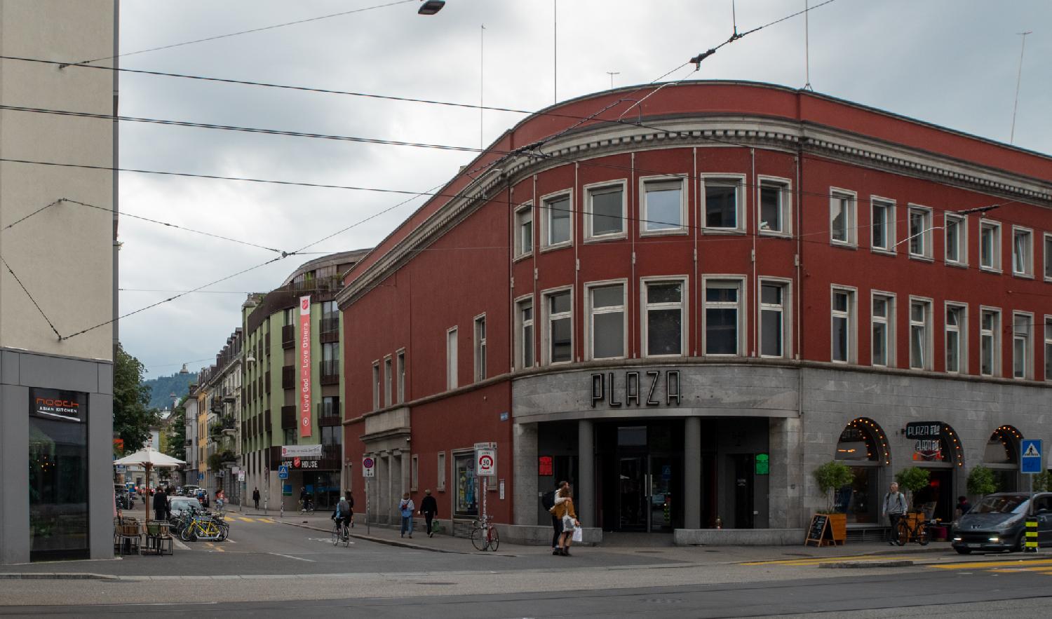 A large building at a corner of two streets. The building is red, the corner is round, and there's a PLAZA neon sign on top of the door.