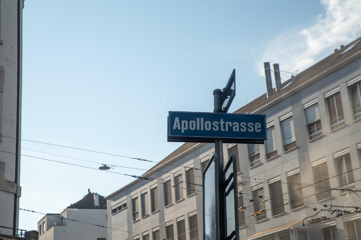 "Apollostrasse" blue street sign in front of tram cables and a building in the background.