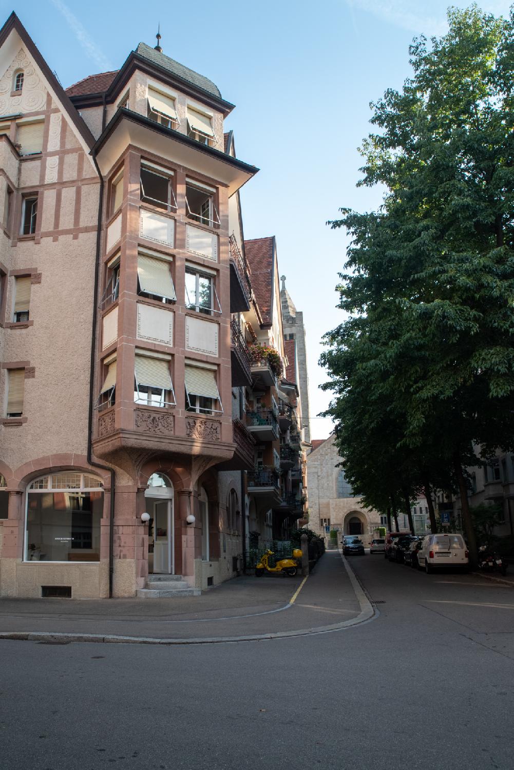 A light brown-red building on a street corner, with rectangular bay windows topped by a cloister vault roof and arched doors and windows on the ground floor. In the far end of the picture, a part of a white church is visible.