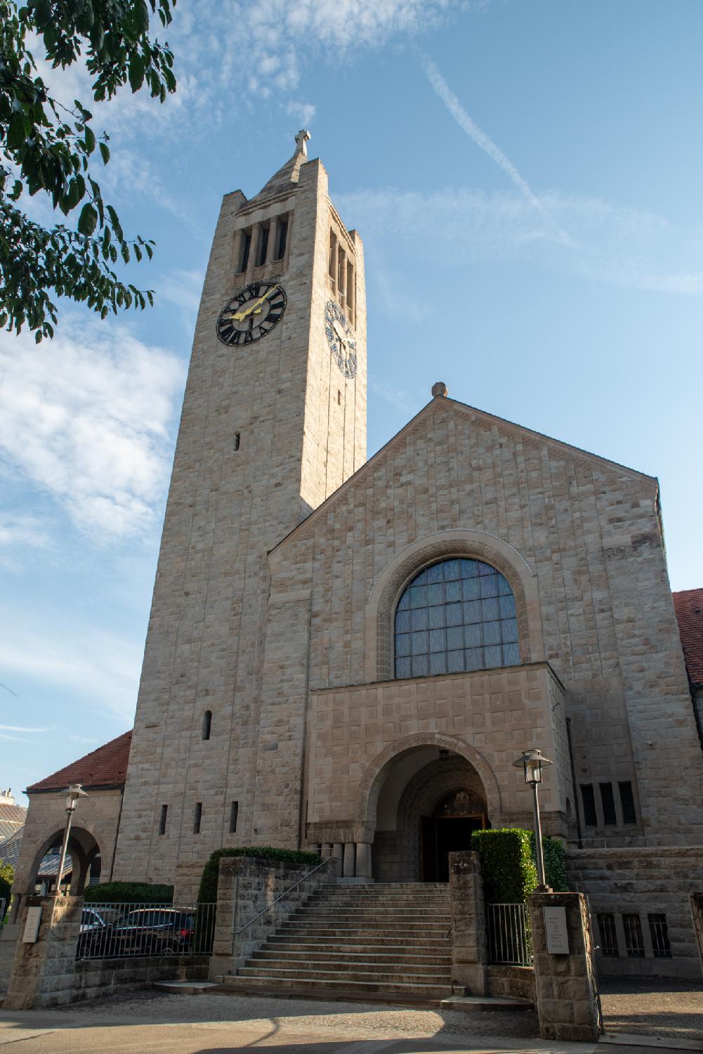 A sandstone church with a church tower topped by tented roof and a cross. The entrance is a wide staircase ending on a round arched door; there's a round arched window on top of it.