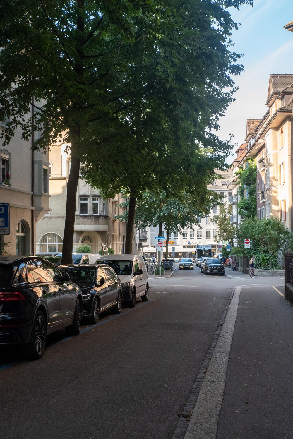 A narrow street in a city between 4-5 story buildings, with cars parked under trees on the left side.