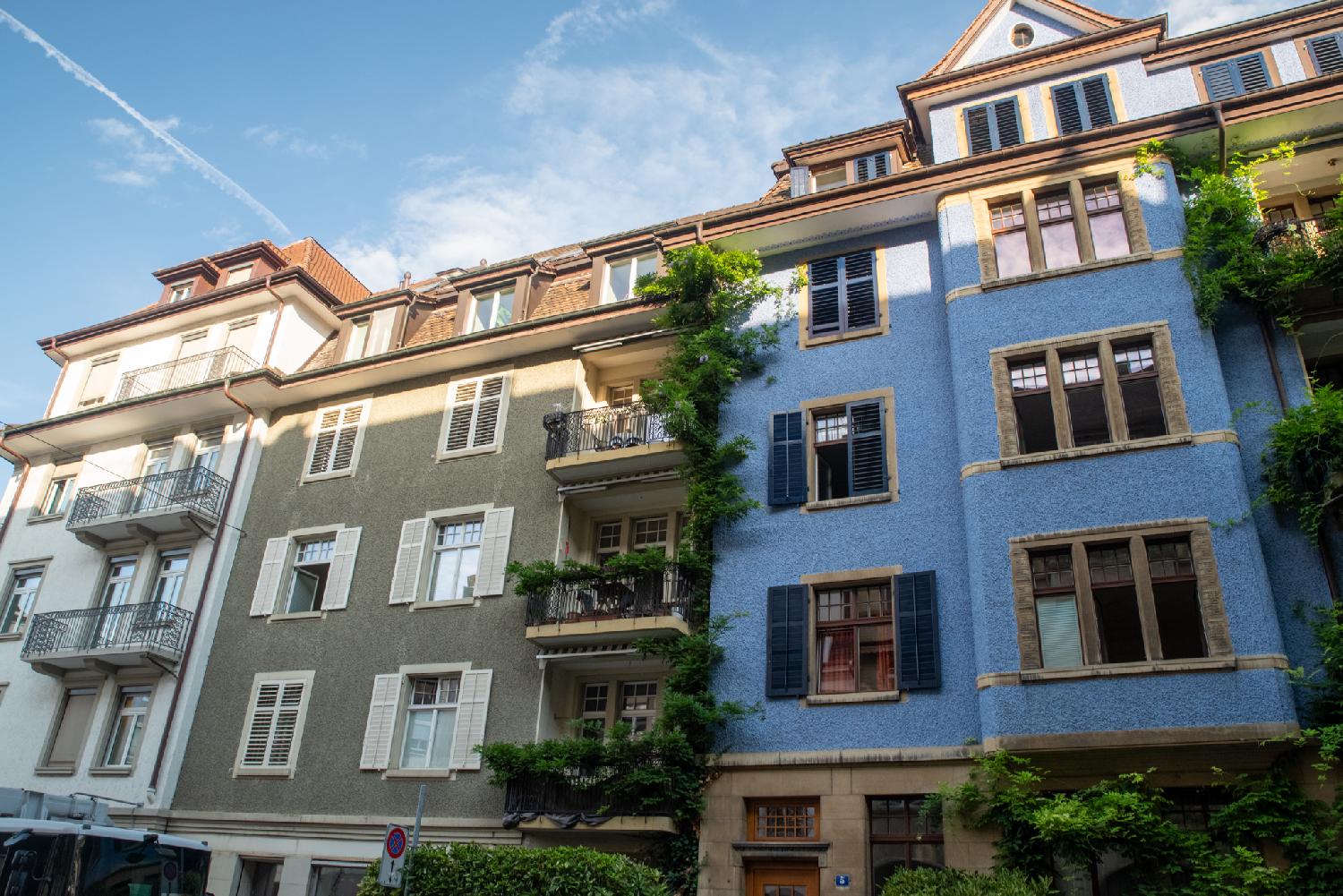 A white, green-brown and a blue facade of 5-story buildings  with balconies and vegetation
