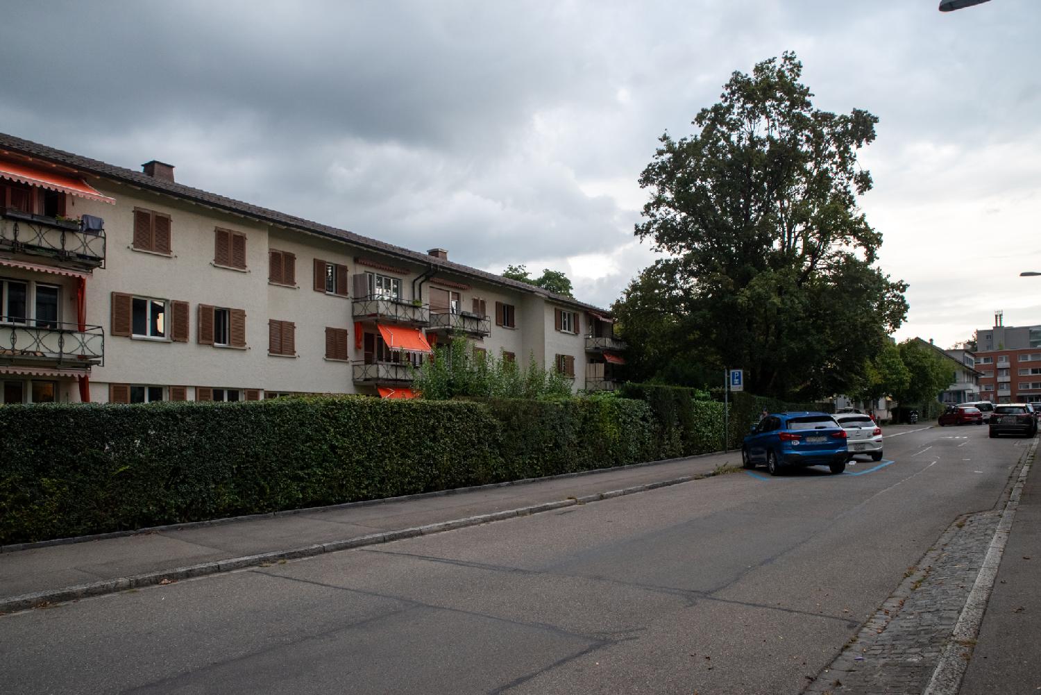 A residential street with 3-storey beige buildings with brown blinds behind a hedge