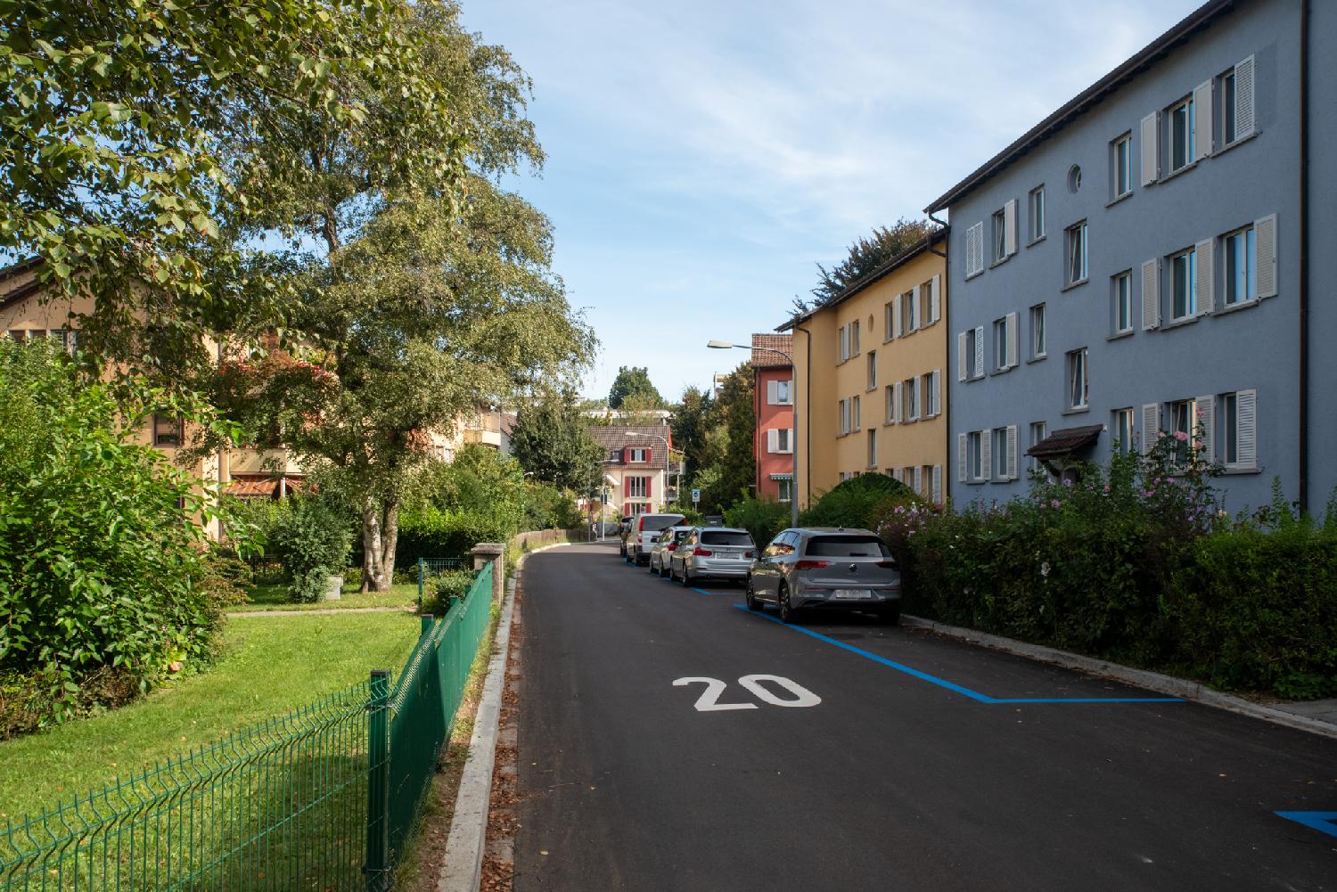 A residential street with a garden on the left and 3-story colorful buildings on the right, with cars parked in blue parking spot and a "20" painted on the street.