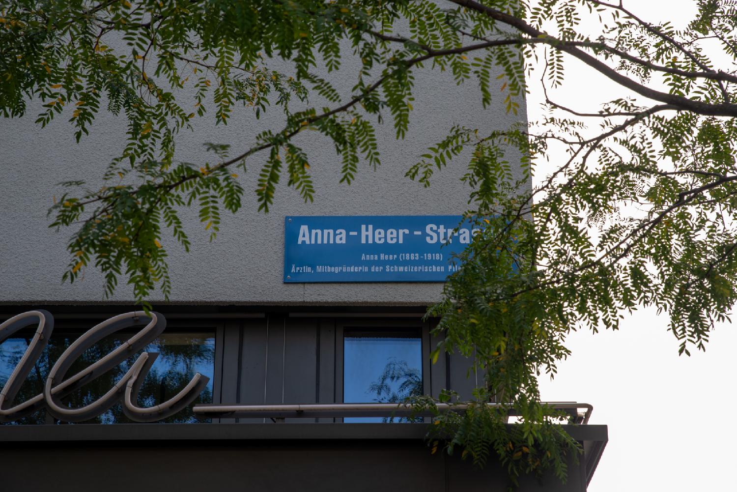 A blue street name sign, partly hidden by a tree in front of it. The text reads "Anna-Heer-Strasse - Anna Heer (1863-1918) - Ärtztin, Mitbegründerin der Schweizerisches Pflegerinnenschule".