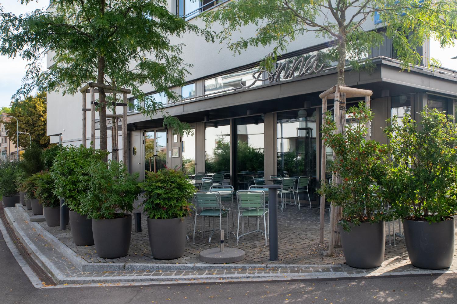 The terrasse of a closed café, with green chairs and potted plants protecting it from the street. The café is called Anna.