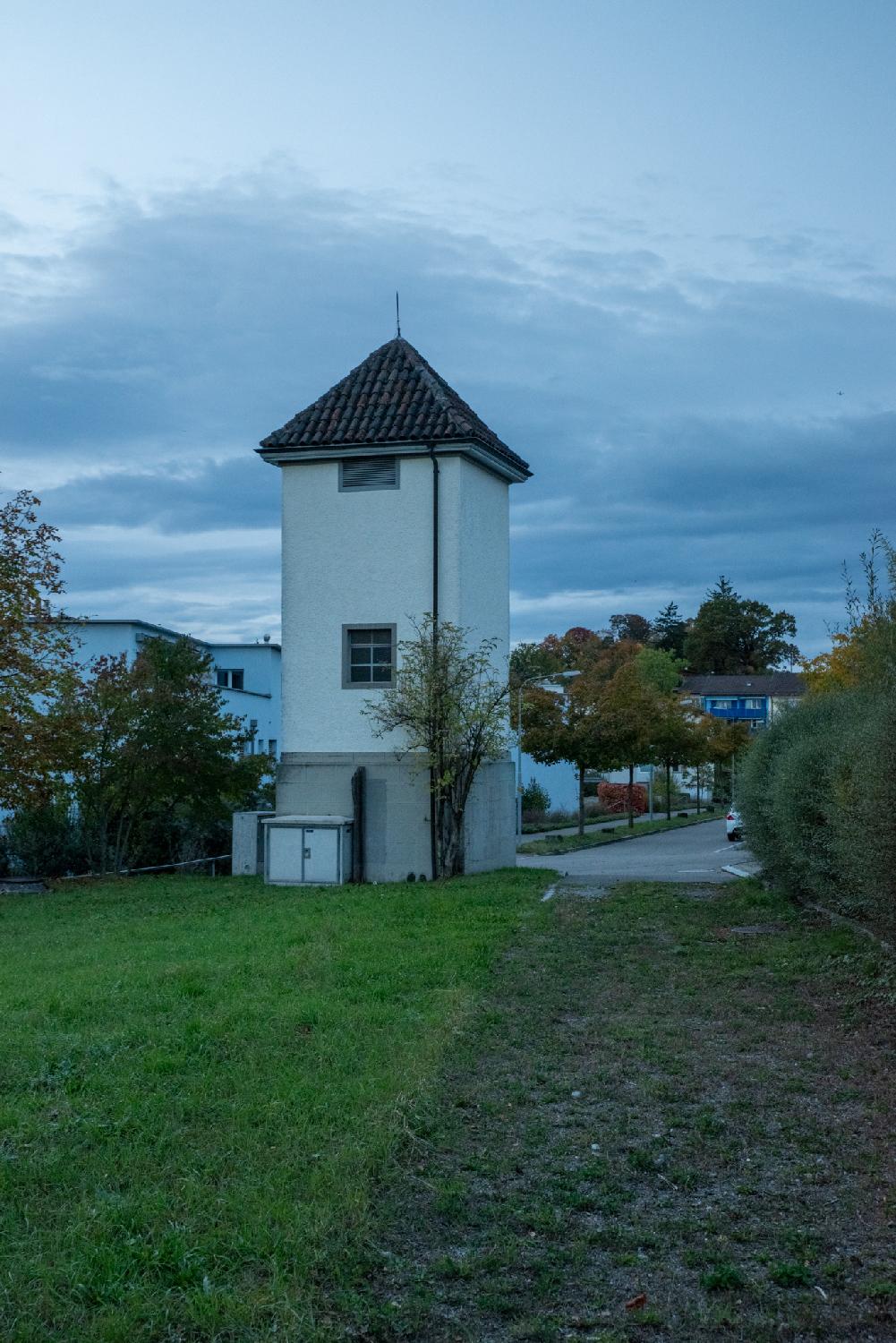 A white 3-storey tower with a pyramid roof, next to a dirt path on the edge of a road