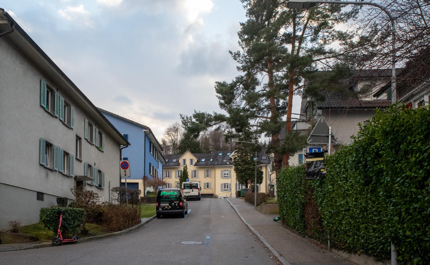 A residential street with 3-story buildings and a couple of vans parked on the left side of the street and hedges on the right side.