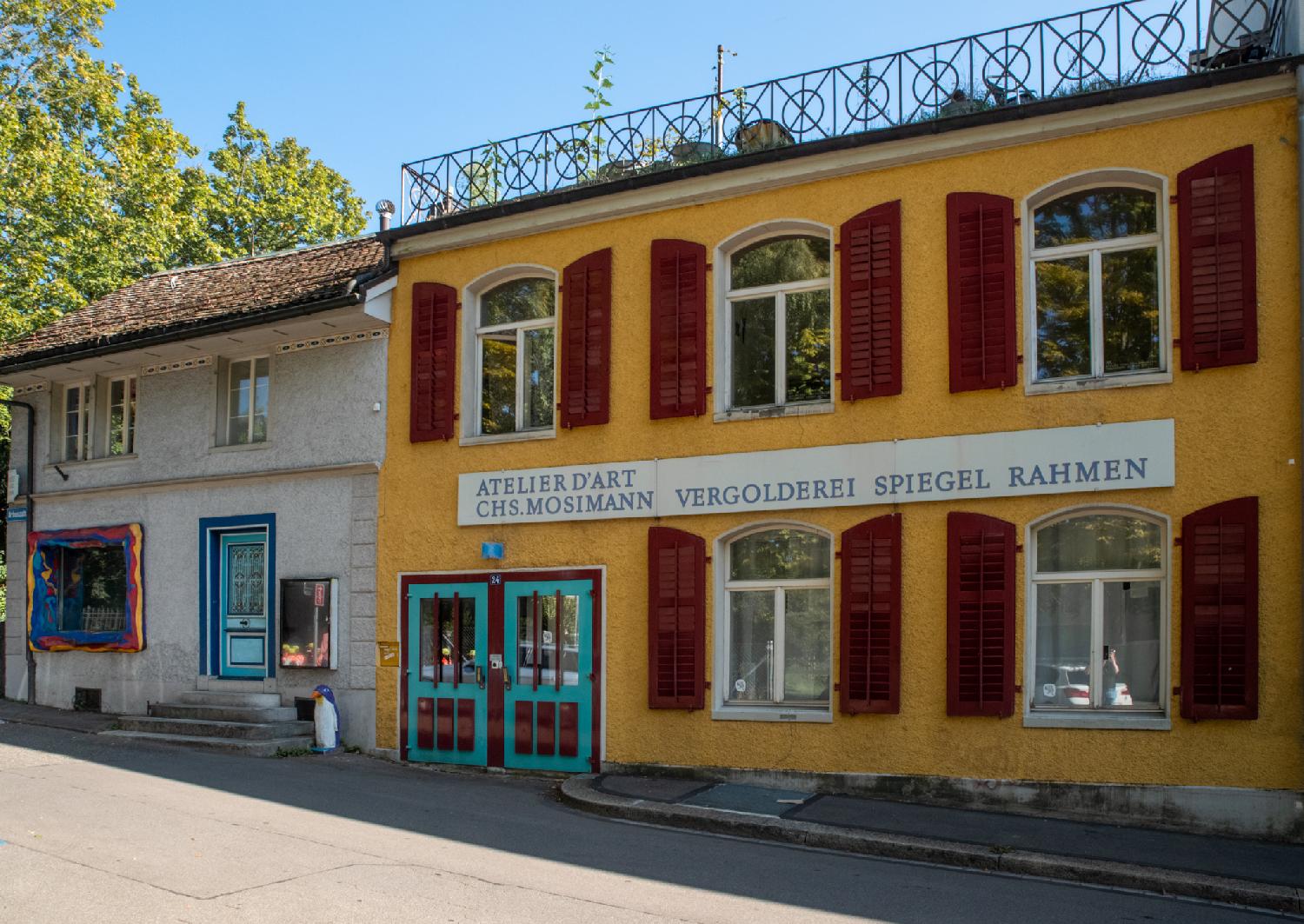 Two-story buildings at the end of a street. The right building has a roof terrace and is bright yellow with dark red blinds and a turquoise double door. The sign on the building reads "Atelier D'Art, CHS. Mosimann, Vergolderei Spieguel Rahmen". The left side of the building has a very colorful window, a turquoise door with a few steps in front of them, and a penguin statue.